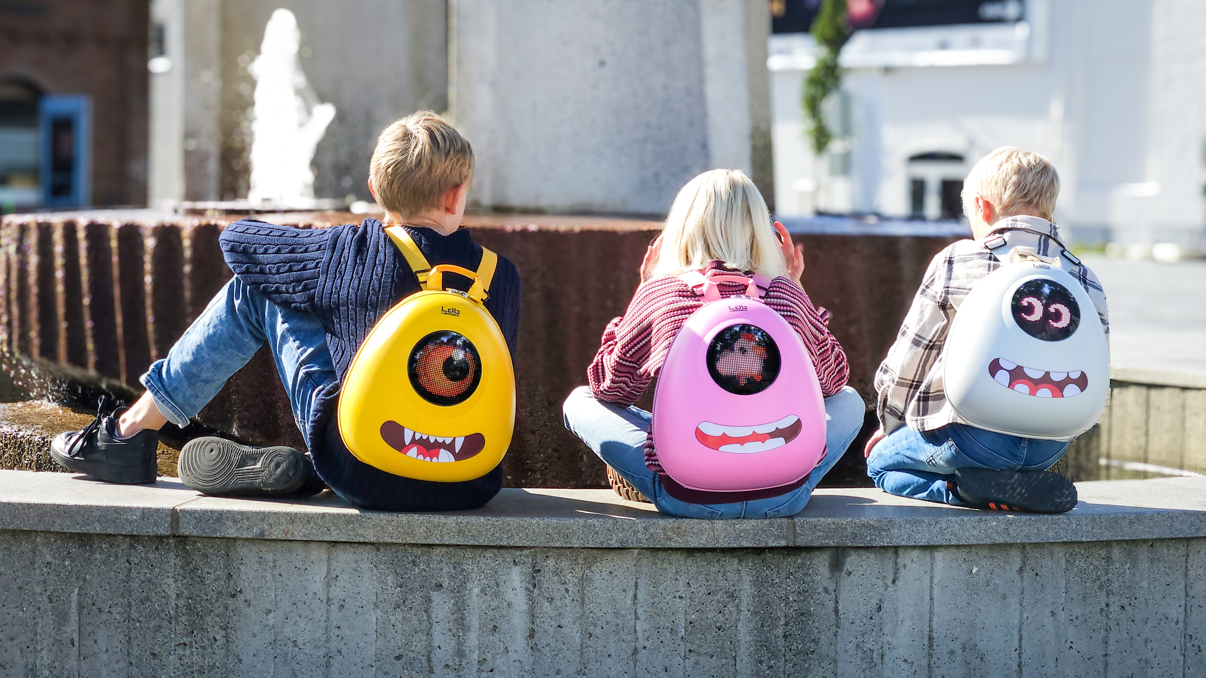 Three kids sitting on a bench by a fountain wearing Ledback Buddy backpacks in Yellow, pink and White, with eye design LED screens and teeth funny stickers.