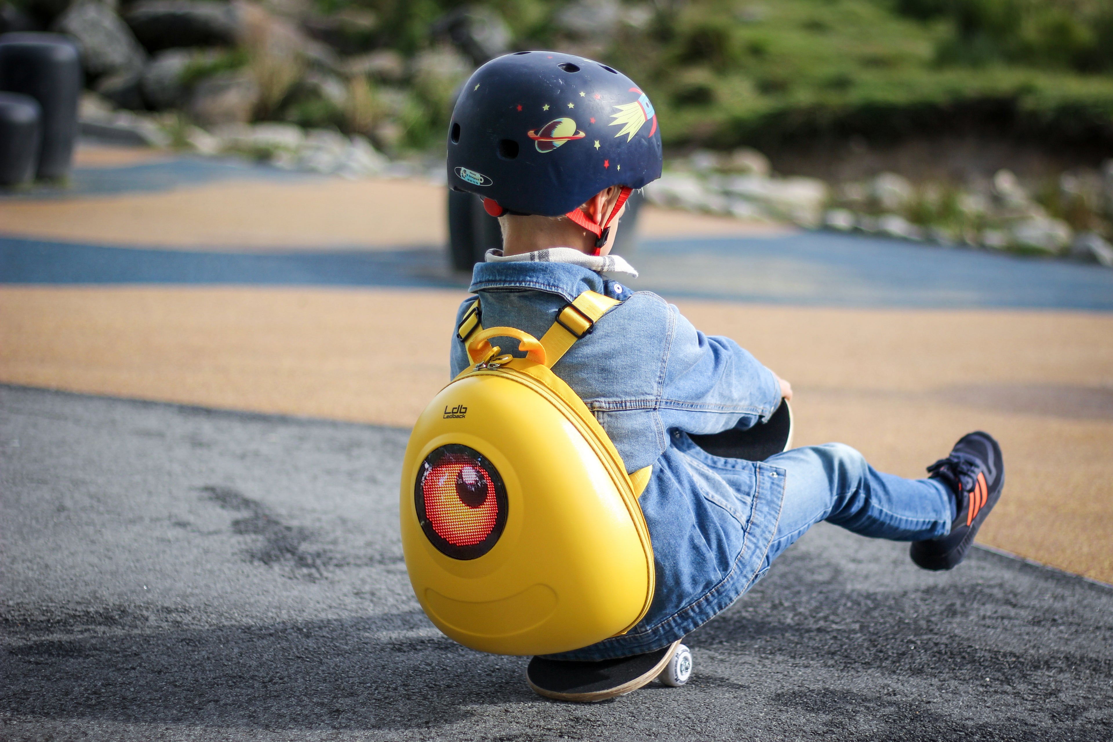 Small boy in denim sitting on a skateboard wearing a blue helmet with rockets, and a Lemon Drop Yellow hard shell backpack from Ledback featuring a circular LED screen displaying an orange eye.