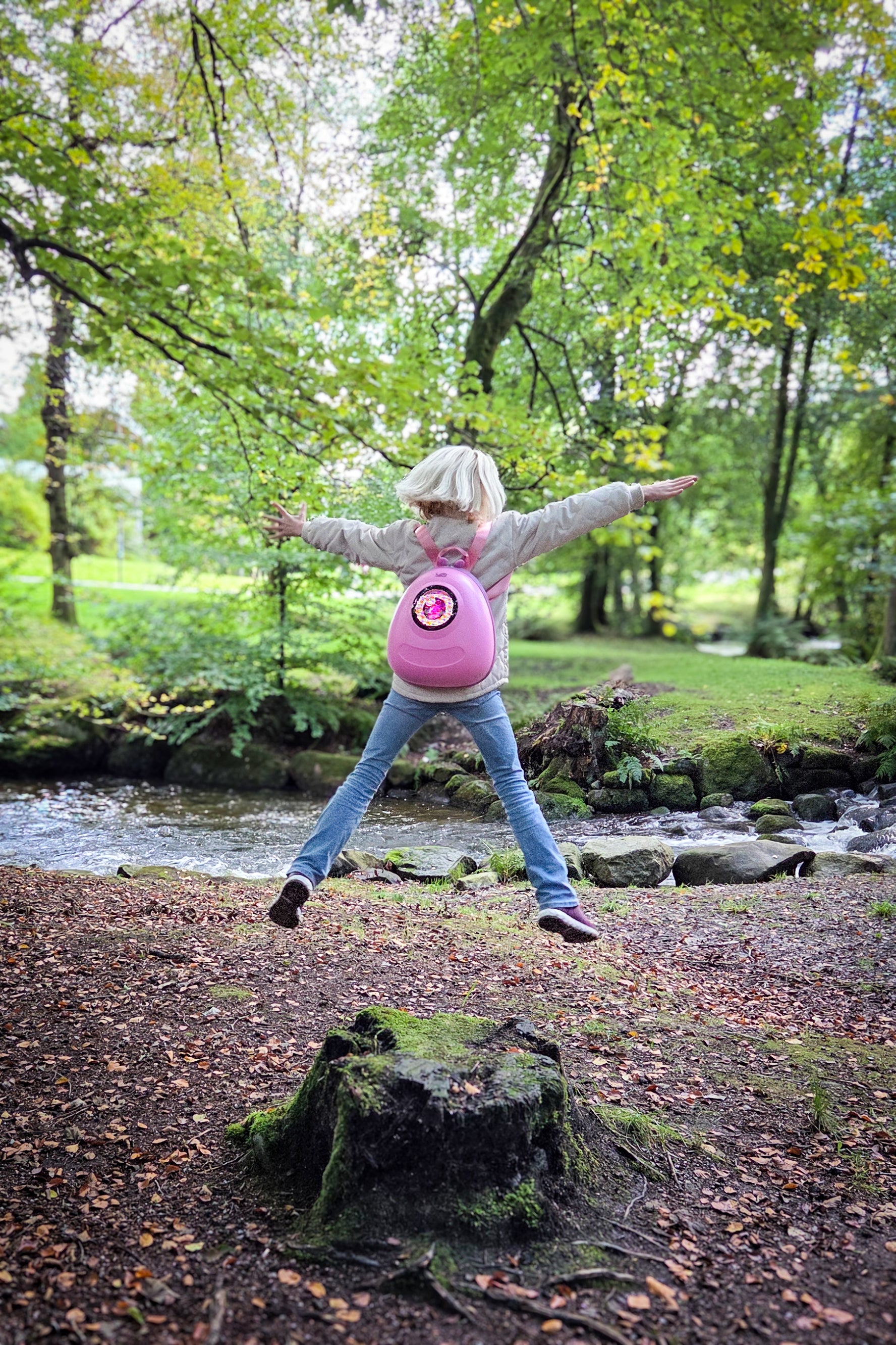 Girl jumping like a starfish off a tree stump in a park, while wearing a Raspberry Sorbet Pink hard shell backpack from Ledback, with a round pink and white eye lighting up on a round LED screen.