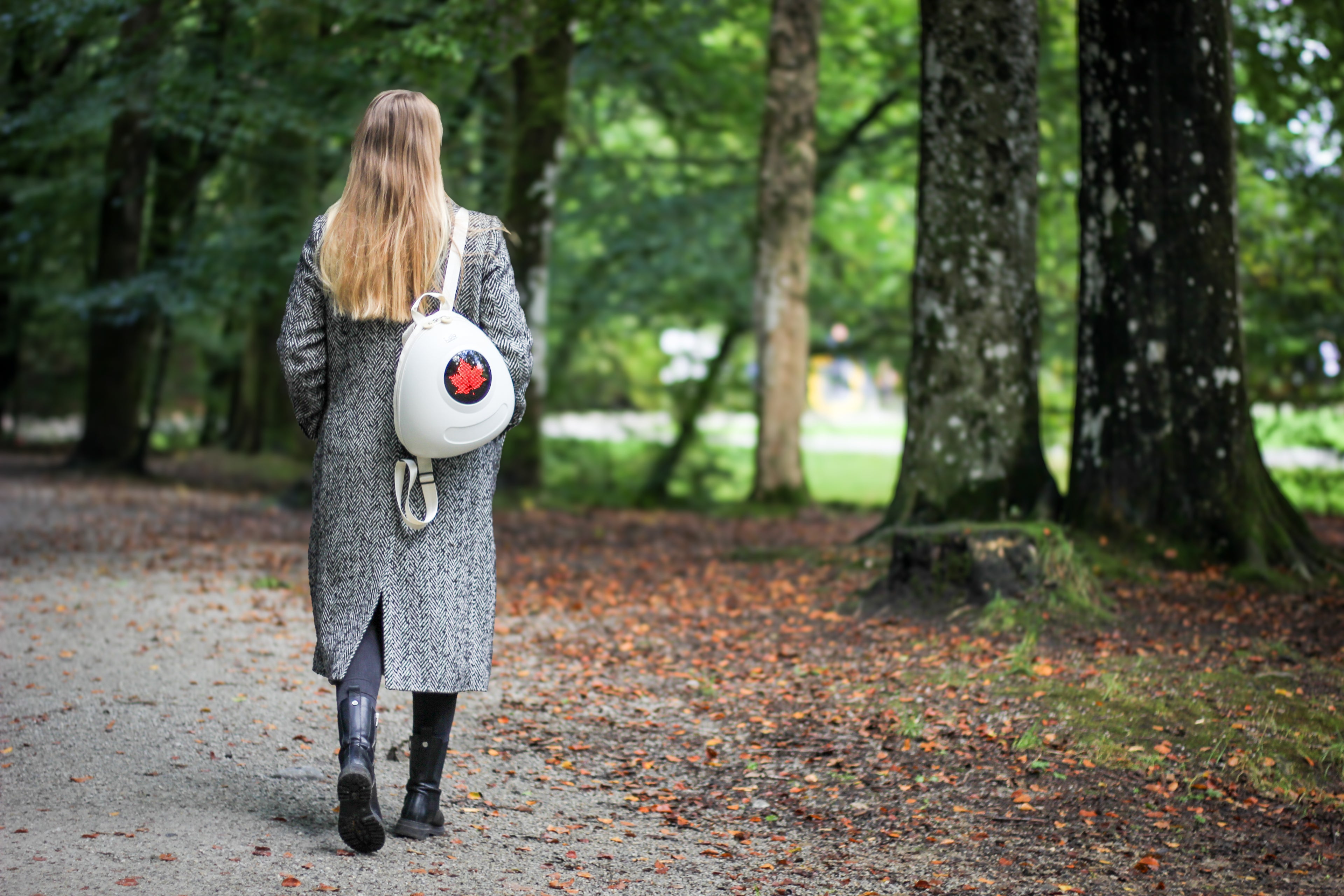 Lady with long hair in a grey coat walking through park wearing a Whipped Cream White Ledback Buddy backpack featuring an LED screen displaying a red maple leaf.