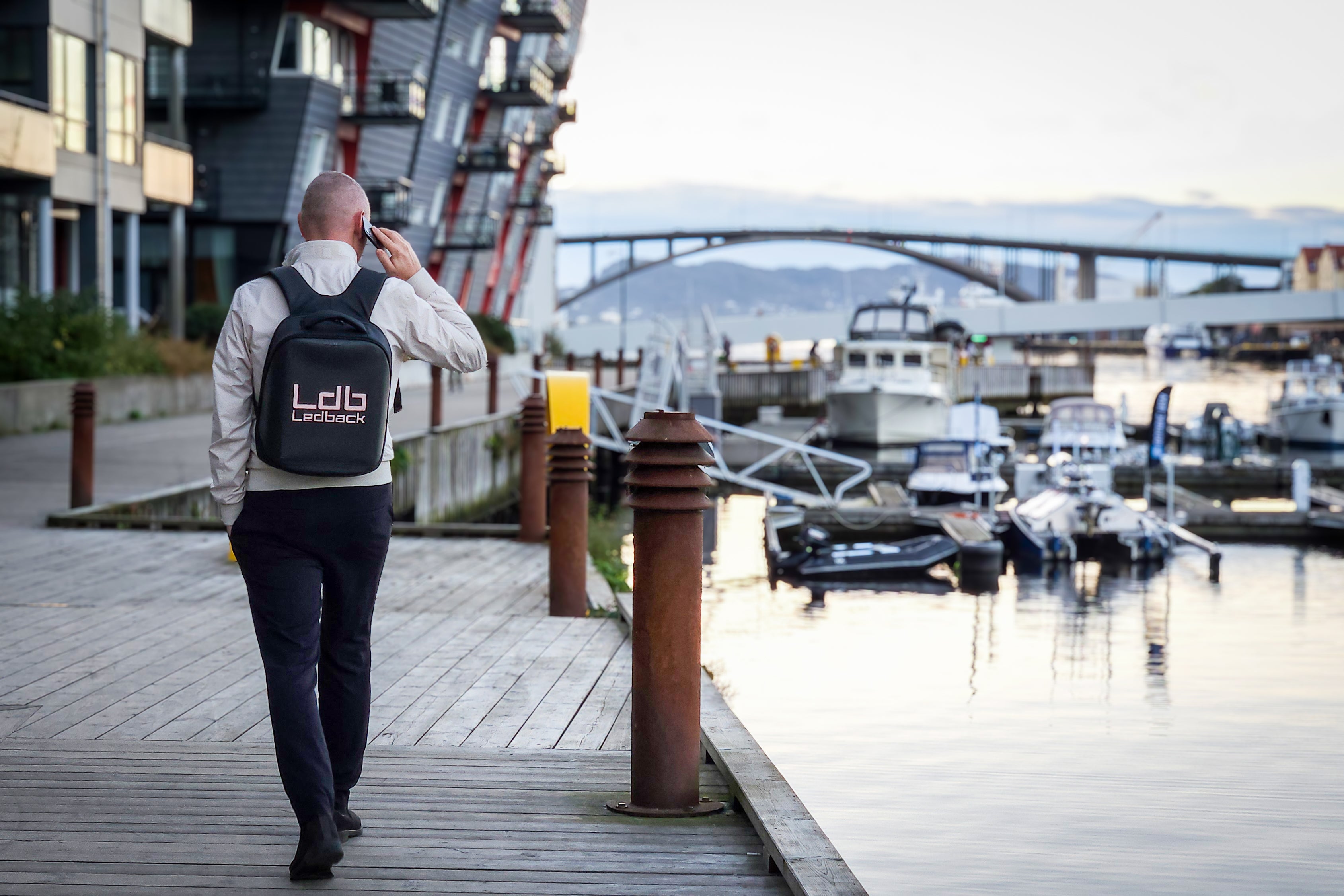 Man in elegant clothes, walking along the docks of Bergen, wearing a Black Ldb Ledback HD Smart LED Laptop Backpack with the LED screen displaying the logo of LDB Ledback, with modern houses, boats and a bridge in the background